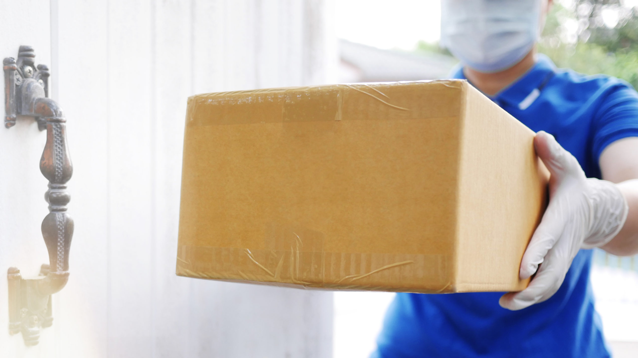 Delivery services courier during the Coronavirus (COVID-19) pandemic, close-up of cardboard box holding by a courier wearing protective face mask and latex gloves at home front door blurred background